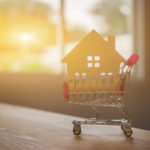 A cutout of a house in a tiny shopping cart on a table in front of a window with a sunset.